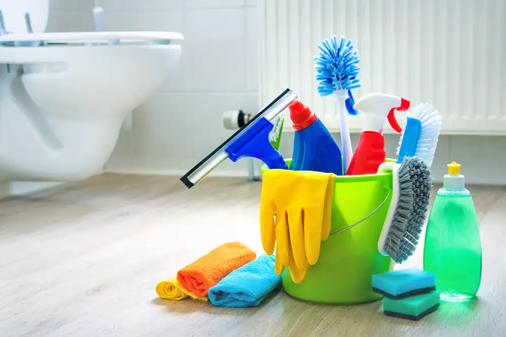 A green bucket filled with cleaning supplies sits on a bathroom floor near a toilet&mdash;perfect for tackling chores or highlighting top-notch cleaning services in Camden and Atlantic County, NJ. Sponges, dish soap, and towels are neatly arranged nearby.