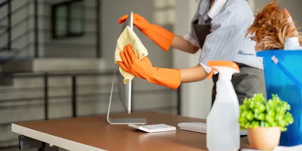 A person wearing orange gloves cleans a computer monitor with a cloth on a desk. Nearby are a keyboard, spray bottle, feather duster, blue cleaning bucket, and a small potted plant—perfect for NJ cleaning services Camden and Atlantic County.